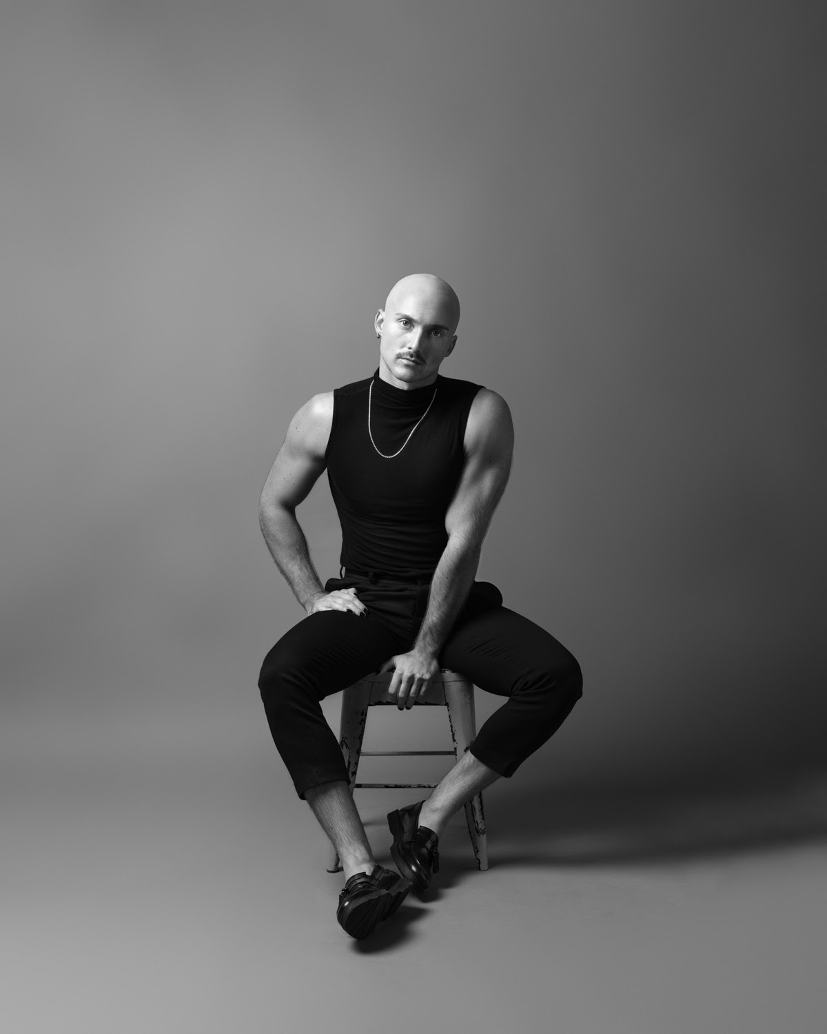 Black and white photo of J. Logan, a white person with a shaved head, sitting on a stool and facing the camera with a serious expression. They are wearing a black, sleeveless turtleneck, black pants, and blank shoes.