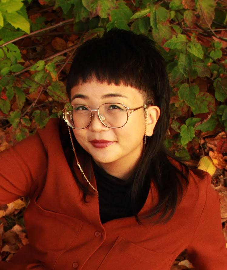 A Korean-American woman with large glasses and black turtleneck smiles at the camera. She is in 3/4 profile and there is autumn foliage in the background.